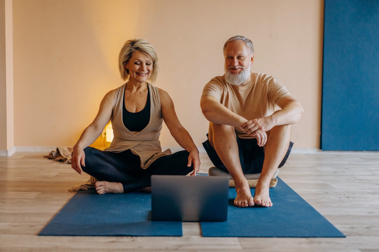 about-img-01 An elderly couple practicing yoga at home with online guidance, smiling and relaxed.