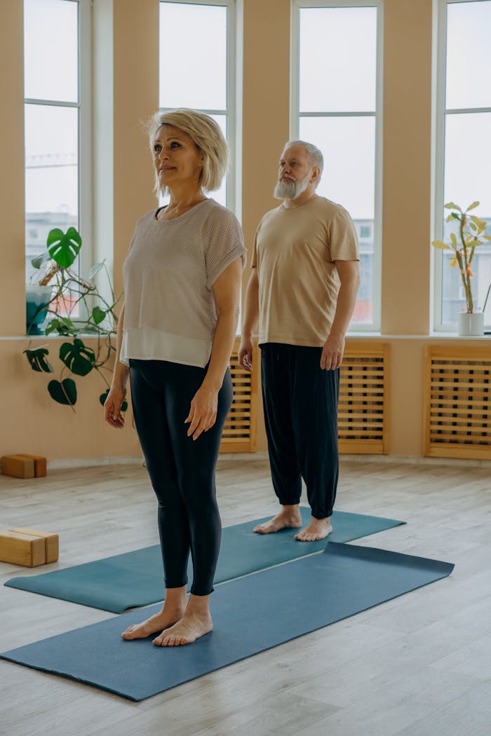 gallery-02 Elderly man and woman in yoga pose on mats in a sunlit indoor studio, embodying healthy lifestyle.