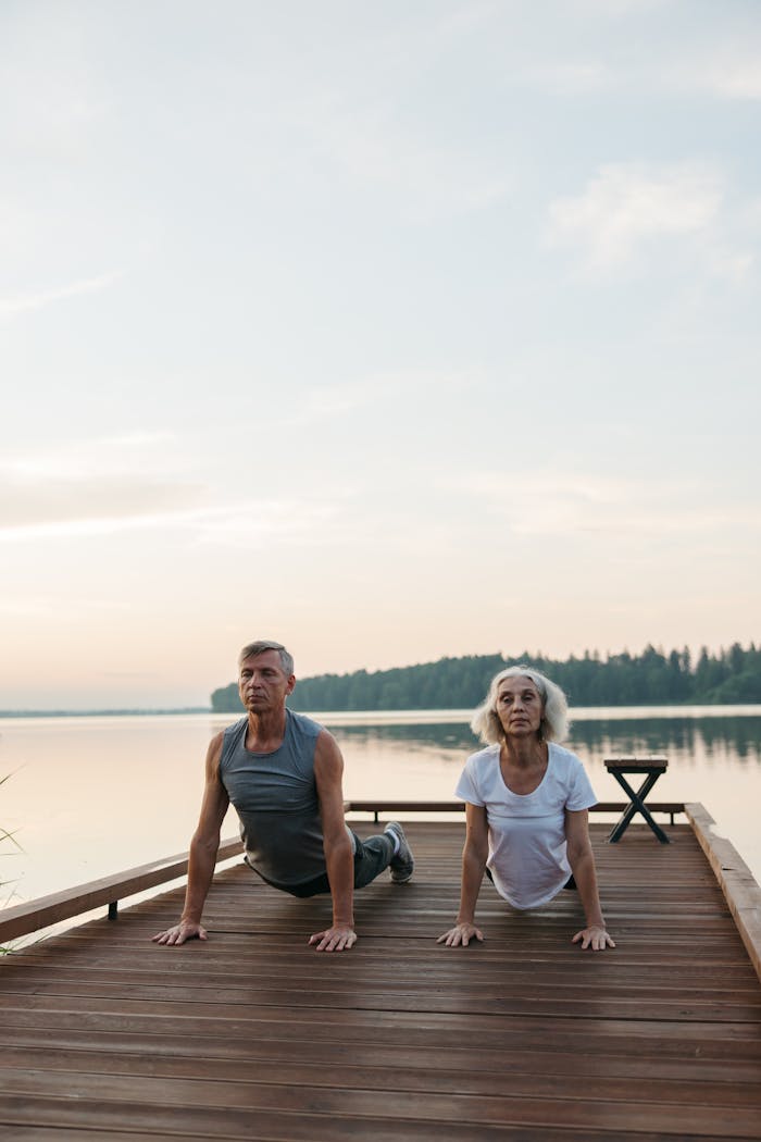 gallery-05 Elderly couple performing yoga outdoors by a serene lake.